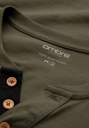 Klassisk henley-tröja i olivgrön bomull med rund halsringning, svart knappslå och träknappar. Etiketten anger 100% ren bomull, storlek M.