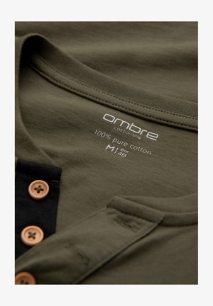 Katoenen henley shirt in olijfgroen met een ronde halslijn, zwarte bies en houten knopen. Etiket geeft 100% puur katoen aan, maat M.