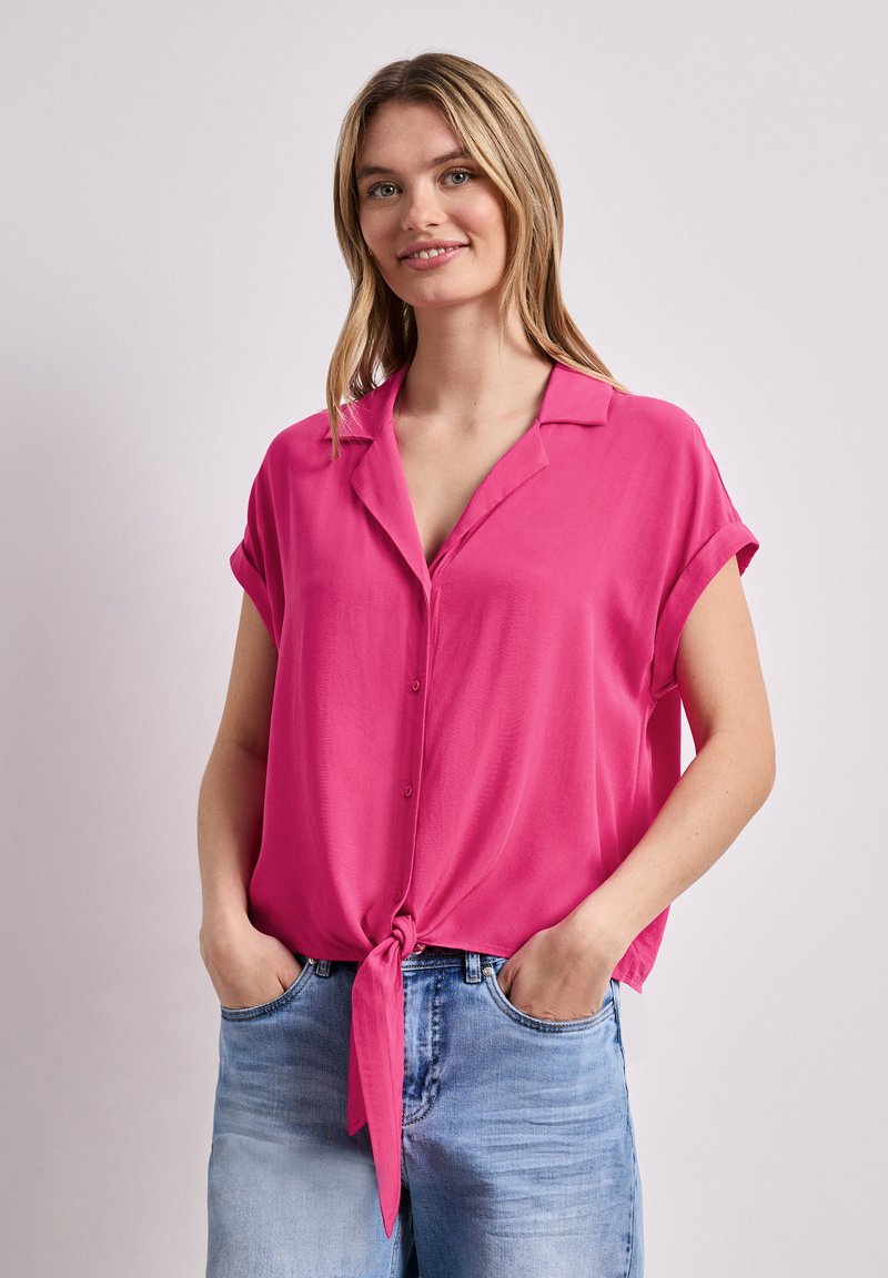 Jeune femme aux cheveux blonds porte une chemise rose vif à manches courtes nouée à la taille et un jean bleu clair, les mains dans les poches.