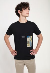 Sort bomulds T-shirt med en tekstureret grafisk lommeprint, der har en landskabsillustration, suppleret med hvid tekst på grafikken.