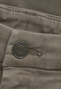 Bruine stof met fijne textuur, voorzien van een metalen knoop met gegraveerd logo en topsteekdetails langs de naad.