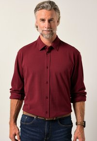 Camicia button-up bordeaux con maniche lunghe, realizzata in tessuto morbido. Presenta un colletto classico e un sottile dettaglio dei bottoni. Indossata con jeans in denim.