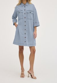 Robe chemise en denim bleu clair avec des manches courtes, deux poches avant et fermeture par boutons. Présente une coupe décontractée et une longueur jusqu'aux genoux.