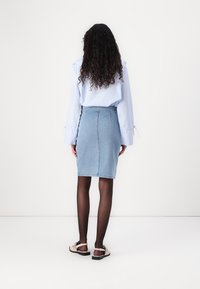 Jupe crayon en denim bleu clair avec une taille haute, texture lisse et détail de couture au dos, associée à une chemise rayée bleue et des collants noirs.