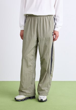 Persoon die licht olijfgroene losse broek draagt met zwarte zijstrepen, witte top met lange mouwen en witte sneakers op een groene vloer.