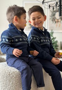 Kaksi poikaa istuu tekstuurilla varustetulla valkoisella pinnalla, pukeutuneina tummansinihin neulepaitoihin, joissa on kuvioita, ja tummiin housuihin, pitäen käsissään välipaloja.