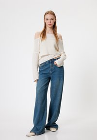 Lichte, off-shoulder, gebreide trui in crème, gecombineerd met een broek van blauwe denim met wijde pijpen. Model poseert tegen een effen witte achtergrond.