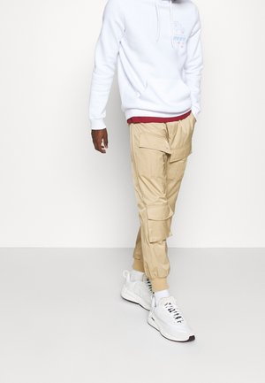 Beige cargo kalhoty s mnoha kapsami, spárované s bílou mikinou s grafickým designem, a bílé sportovní tenisky s černými detaily.