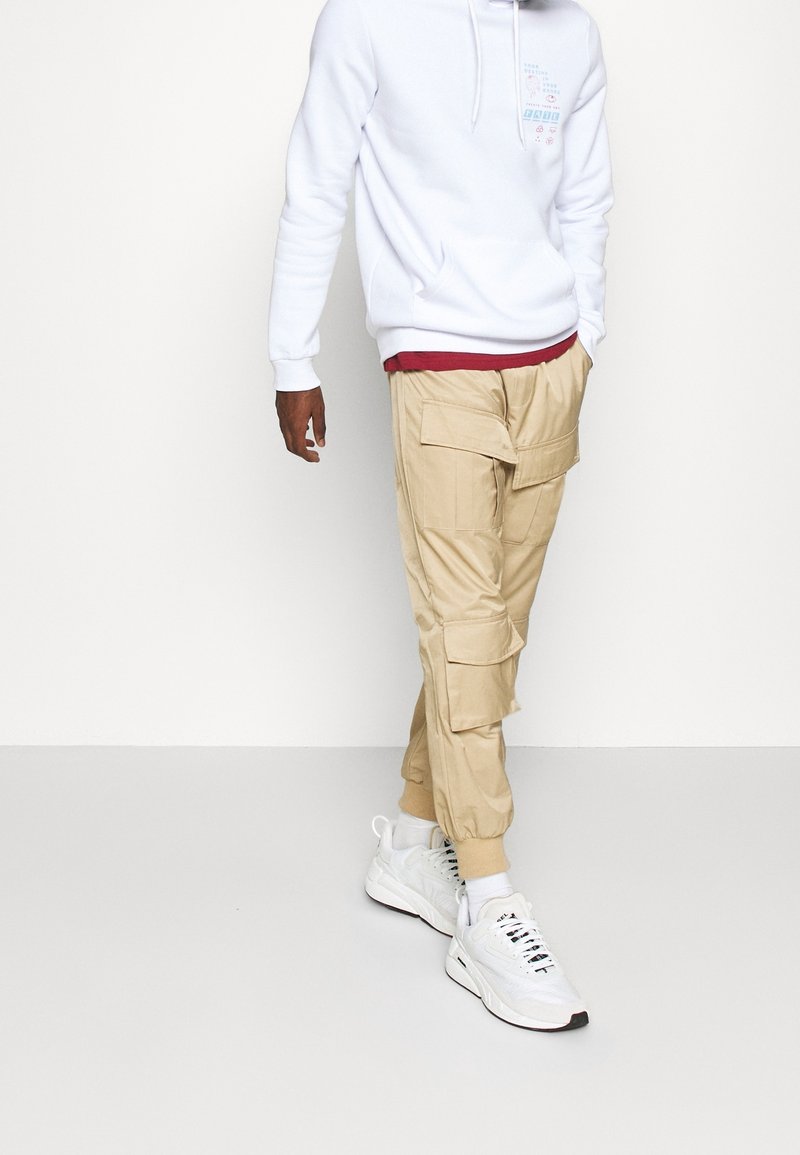 Pantaloni cargo beige con molte tasche, abbinati a una felpa bianca con un design grafico, e sneakers atletiche bianche con accenti neri.