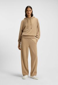 Felpa beige con cappuccio e cordino, abbinata a pantaloni larghi coordinati. Caratterizzata da un tessuto morbido con una consistenza liscia e sneakers bianche.