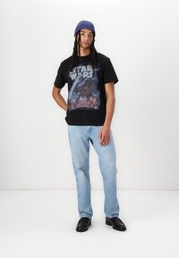 Černé grafické tričko s barevným designem Star Wars, spárované se světlými modrými rovnými džíny a černými nazouvacími botami. Modrý pletený čepice.