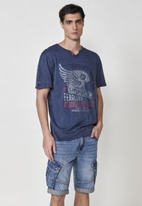 Temna modra kratka majica s kratkimi rokavi in grafičnim tiskom, V-izrezom in gumbnimi detajli, v kombinaciji z modrimi jeans kratkimi hlačami s stranskimi žepi.