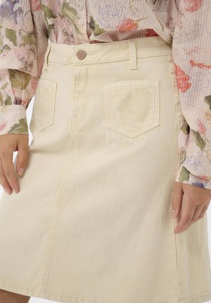 Femme portant une jupe en jean crème avec des poches brodées de feuilles et un chemisier à manches longues à fleurs, mains reposant le long du corps.