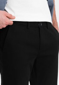 Man som har svarta byxor och en vit skjorta, med hand som greppar midjebandet på vänster sida, beskuren vid midjan och låren.