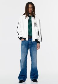 Person med lange dreadlocks iført en hvid lynlåsjakke, grønstribet skjorte, brede blå jeans og sorte sko, stående foran en ensfarvet baggrund.