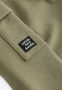 Olivenfarbener Stoff mit einem schwarzen rechteckigen Patch und weißem Text: "Lass es uns jeden Tag angehen." Glatte Textur und lässiges Design.