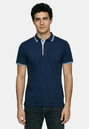 Jeune homme aux cheveux bouclés et foncés portant un polo bleu marine avec un col et des manches rayés blancs, debout devant un fond uni.