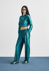 Γυναίκα με μακριά σκούρα μαλλιά και γυαλιά, ντυμένη με τζάκετ αθλητικό της Puma σε τυρκουάζ απόχρωση, παντελόνι φαρδύ ίδιου χρώματος και τυρκουάζ αθλητικά παπούτσια Puma, στέκεται σε μπλε πάτωμα.