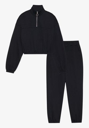 Melns saīsināts sporta krekls ar augstu apkakli un pusapakšu rāvējslēdzēju, kopā ar melnām sporta biksēm ar elastīgu jostas vietu un aprocēm.