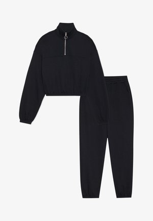 Melns saīsināts sporta krekls ar augstu apkakli un pusapakšu rāvējslēdzēju, kopā ar melnām sporta biksēm ar elastīgu jostas vietu un aprocēm.