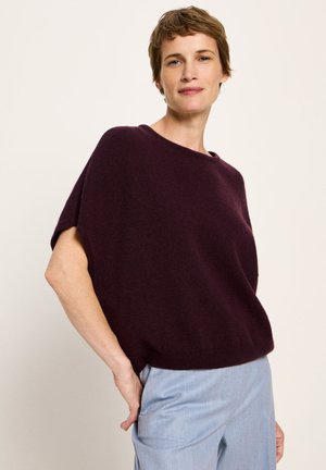 Woman with short brown hair wearing a loose, dark purple short-sleeve sweater and light blue pants, standing against a plain background.
