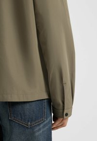 Khaki, let jakke med afslappet pasform. Har manchetter med knap, mat overflade, og er suppleret af mørke denim jeans.