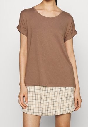 Kobieta nosząca brązową koszulkę z krótkim rękawem i podwiniętymi mankietami oraz beżową spódnicę w kratę z rozcięciem z przodu.