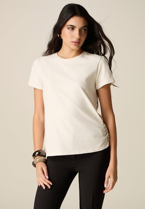 Vrouw met lang donker haar, gekleed in een wit T-shirt met korte mouwen, zwarte broek, gouden oorbellen en dikke armbanden, poserend tegen een neutrale achtergrond.