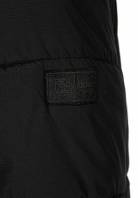 Sort stofjakke med en struktureret overflade. Inkluderer et rektangulært sort mærke med præget logo og teksten "RESPECT".