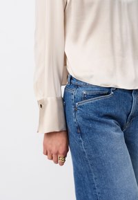 Lichtbeige satijnen blouse met lange mouwen en een manchet met een gouden accent, gecombineerd met blauwe spijkerbroek met een hoge taille.