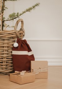 Bordeauxrode katoenen tasje met trekkoord, witte strepen en pompon, naast een gevlochten mand en twee bruine geschenkdoosjes met juten linten.
