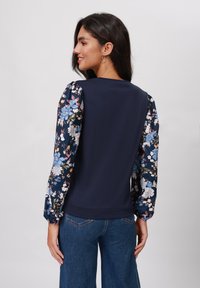 Mørkeblå genser med blomstrete pufferm i blå, hvit og oransje. Glatt tekstur på kroppen, ribbede mansjetter rundt ermene.