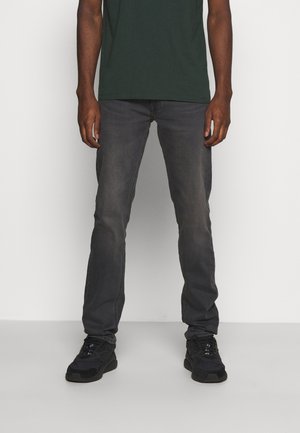Jean gris foncé avec une coupe droite, présentant une finition délavée. Porté avec un t-shirt vert et des baskets noires, mettant en avant un look décontracté.