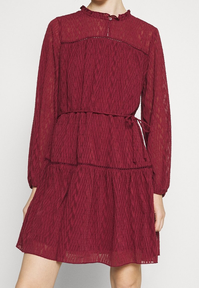 Burgunderrotes, gestuftes Kleid mit langen Ärmeln, Rüschensaum und Kordelzug in der Taille; aus strukturiertem Stoff mit aufwendigen Nähdetails.