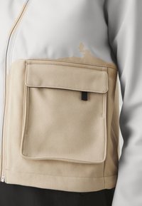 Veste beige et gris clair avec une grande poche avant beige dotée d’un rabat et d’un petit onglet noir, portée sur un pantalon noir.