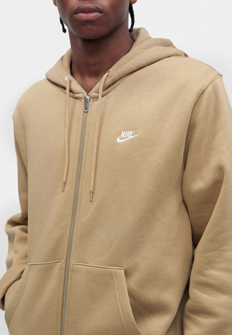 Person wearing a beige Nike zip-up hoodie with hands in front pockets against a plain light background.