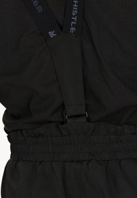 Svarta byxor med elastiskt midjeband och justerbara svarta hängslen med subtilt varumärkestext, bärs över en svart skjorta.