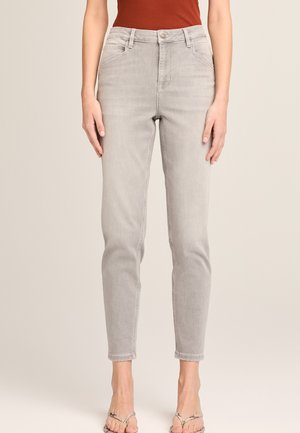 Eng anliegende hellgraue Jeans, getragen von einer Person mit einem rostfarbenen Oberteil, die barfuß vor einem neutralen Hintergrund steht.