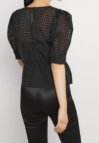 Blusa nera a scacchi con maniche a palloncino, vita sfiancata e dettaglio a chiave sulla schiena. Abbinata a leggings neri aderenti, che mettono in risalto la texture.