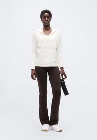 Άτομο που στέκεται φορώντας λευκή κουκούλα, σκούρο καφέ παντελόνι, λευκά αθλητικά παπούτσια, κρατώντας ένα μαύρο παγούρι νερού σε απλό φόντο.