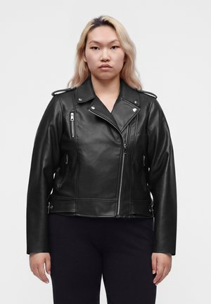 Femme aux longs cheveux blonds portant une veste en cuir noire de motard avec des poches zippées et un pantalon noir, debout devant un fond uni.