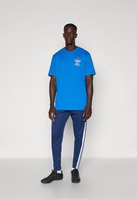 Blue cotton t-shirt with short sleeves, featuring a white graphic logo on the chest. Paired with navy sweatpants and black sneakers.