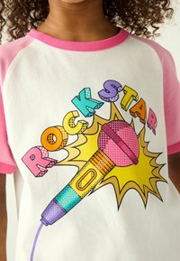 Biely tričko s ružovými raglánovými rukávmi, ktoré zobrazuje farebnú grafiku mikrofónu a text "ROCK STAR" v živých, rozmanitých vzoroch.
