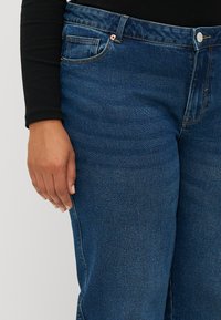 Jeans in denim blu scuro con un fit classico. Le caratteristiche includono cinque tasche, accessori in rame e una leggera usura sulla trama del tessuto.