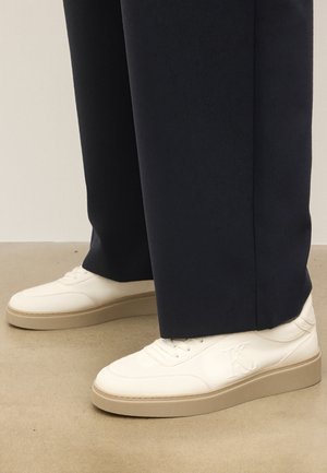 Baskets en cuir blanc avec des semelles beiges et un logo "K." en relief, portées avec un pantalon large noir sur un sol beige.