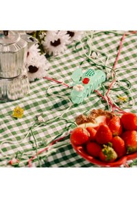 Tissu à carreaux verts et blancs avec des fraises dans un bol rouge, une cafetière en métal, des fleurs de marguerite, une barrette verte et des ficelles décoratives.