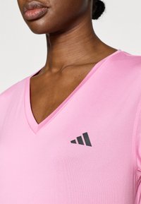 Ružové športové tričko s výstrihom do V, vyrobené z hladkej látky, s čiernym logom na ľavej strane hrudníka a krátkymi rukávmi.
