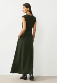 Maxi sukienka w ciemnozielonym kolorze; dopasowany, teksturowany gorset z krótkimi rękawkami, zwiewna spódnica i boczne kieszenie. Noszona z czarnymi botkami za kostkę.