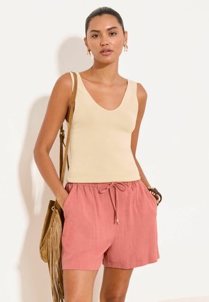 Kvinde iført beige ærmeløs top, lyserøde shorts med snøre, gyldne creoløreringe og bærer en brun skuldertaske med frynser.