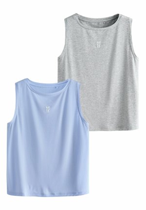 Junge Frau mit lockigem Haar, die ein ärmelloses graues Oberteil mit dem Schriftzug "self" trägt, steht mit den Händen in der Hüfte und schaut seitlich vor einem schlichten Hintergrund.